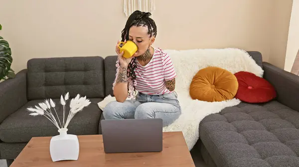 Hispanic amputee woman, seriously focused, utilizing laptop at home; sitting on sofa, sipping coffee, tattoos tracing her armless side.