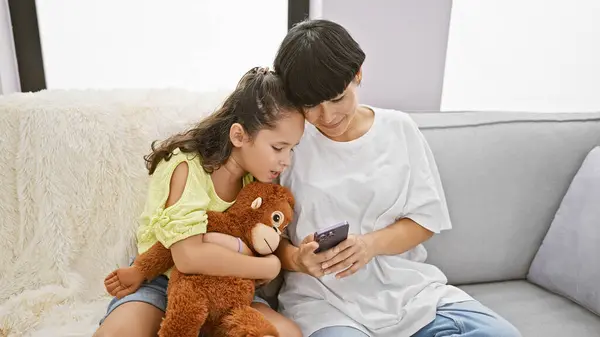 Casual fun at home, mother and daughter enjoy sitting on the sofa, hugging their lovely monkey doll while texting on their smartphone