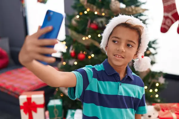 自宅でクリスマスツリーで立っているスマートフォンでセルフィーを作る愛らしいヒスパニックの少年 — ストック写真