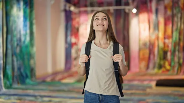 Young beautiful girl student wearing backpack looking around smiling at art gallery
