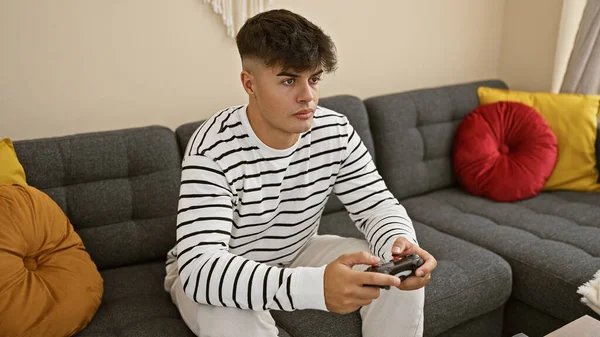 Handsome young hispanic man comfortably sitting on his living room sofa, lost in a serious gaming session. he\'s using a gamepad, fully immersed in online gaming at his cozy home apartment.