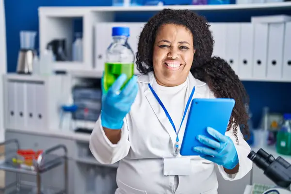 African american woman scientist holding bottle using touchpad at laboratory