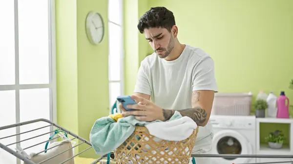 Mladý Hispánec Muž Pomocí Smartphone Opírá Koš Oblečením Prádelně — Stock fotografie