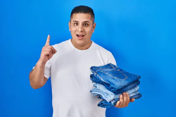 Jovem Hispânico Segurando Pilha Jeans Dobrados Surpreso Com Uma Ideia — Fotografia de Stock