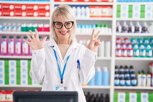 Young caucasian woman working at pharmacy drugstore showing and pointing up with fingers number seven while smiling confident and happy.