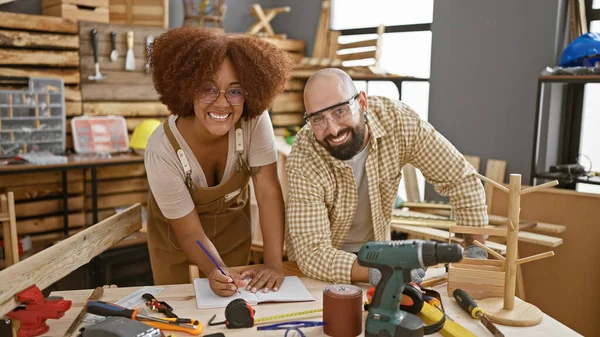 Two smiling carpenters confidently taking notes in their woodworking workshop