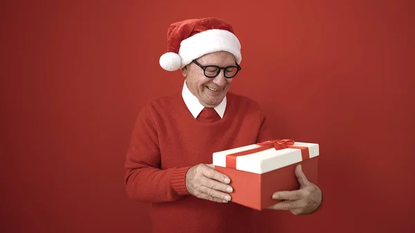 Senior Dragen Kerstmuts Uitpakken Geschenk Geïsoleerde Rode Achtergrond — Stockfoto