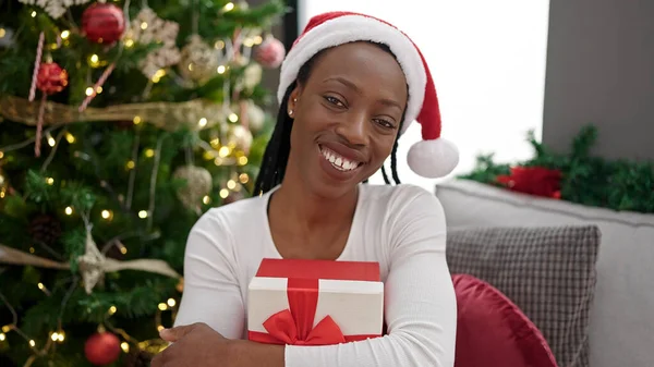 African american woman holding gift sitting on sofa by christmas tree at home