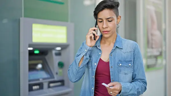 Young beautiful hispanic woman talking on smartphone holding credit card at bank teller