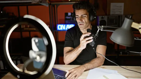 Young hispanic man speaking in a radio show recording video by smartphone at radio studio