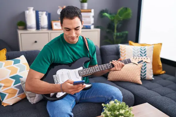 Young hispanic man playing electrical guitar using smartphone at home