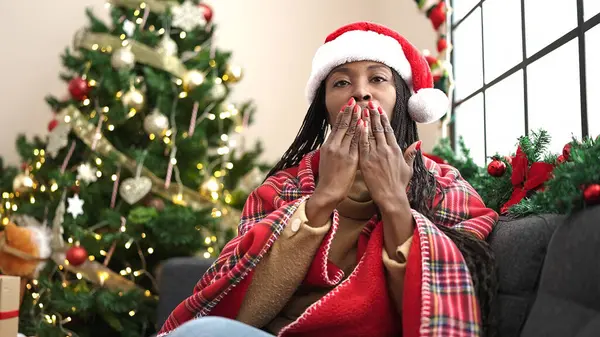 African woman sitting on sofa by christmas tree blowing kissing at home