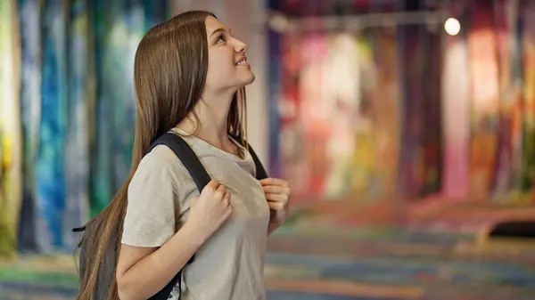 Young beautiful girl student wearing backpack looking around smiling at art gallery