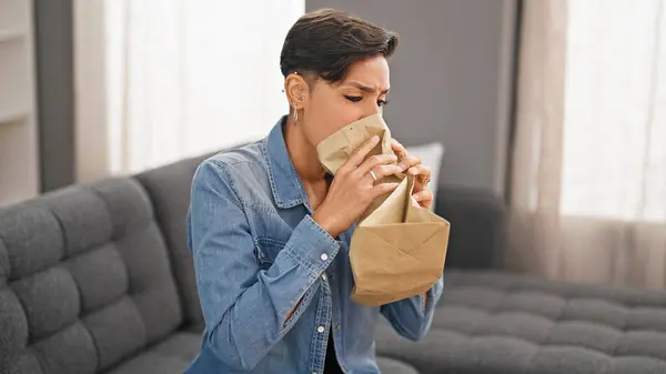 Young beautiful hispanic woman stressed sitting on sofa blowing air on paper bag at home