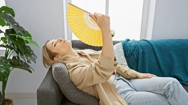 Stunning young blonde woman suffering in hot weather indoors, desperately fanning herself with a hand fan in her cosy home living room