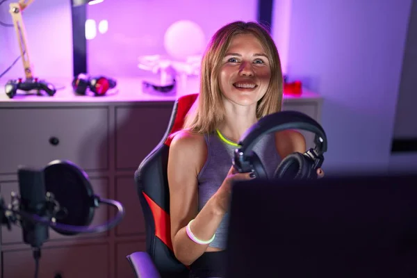 Young blonde woman streamer playing video game holding headphones at gaming room
