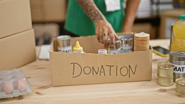 Tattooed, hispanic man\'s gracious hands volunteer, putting canned food donations in a cardboard box at a bustling charity center - the beautiful side of working in altruism and unity