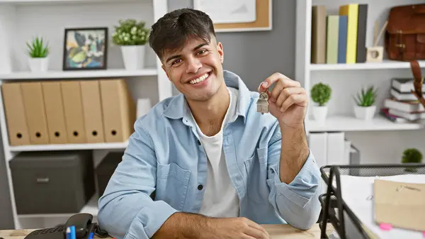 Smiling young hispanic real state agent in the office, celebrating success as he holds the key to a new house - the symbol of his rewarding job.