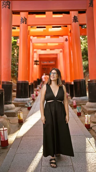 Confident hispanic beauty in glasses, grinning ear-to-ear, looking around in awe at orange-red torii tunnel at fushimi temple in kyoto!