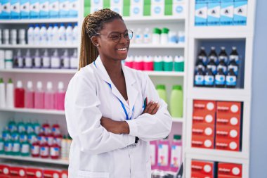 Afro-Amerikalı kadın eczacı gülümseyerek eczanede kollarını kavuşturup duruyor.