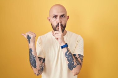 Sarı arka planda dikilen İspanyol dövmeli adam sessiz olmak istiyor. Parmakları dudaklarında, elleri yana doğru işaret ediyor. sessizlik ve gizli konsept. 
