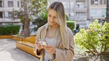 Çekici genç sarışın kadın telefonunu mutlu bir şekilde kullanıyor, dijital bir sohbete dalmış ve yeşil şehir parkında neşeli bir şekilde mesajlaşıyor.