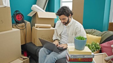 Young hispanic man using laptop sitting on sofa at new home