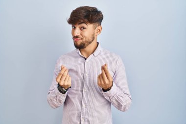 Mavi arka planda duran sakallı bir Arap. Elleriyle para hareketi yapıyor. Maaş istiyor. 