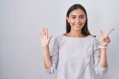 Beyaz arka planda duran genç İspanyol kadın yedinci parmaklarını gösterip işaret ederken kendinden emin ve mutlu bir şekilde gülümsüyor.. 