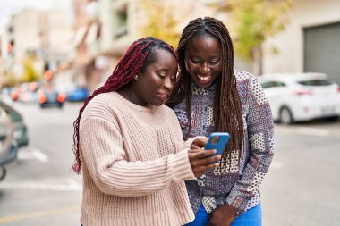 Afro-Amerikalı kadın arkadaşlar, sokakta akıllı telefon kullanarak gülümsüyorlar.