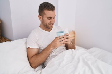 Young caucasian man drinking cup of coffee sitting on bed at bedroom