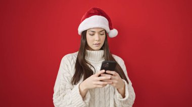 Genç, güzel, İspanyol bir kadın yılbaşı şapkası takıyor ve izole edilmiş kırmızı arka plan üzerinde akıllı telefon kullanıyor.