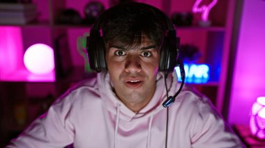 Ciddi bir yüze sahip, odaklanmış genç İspanyol bir adam, karanlık bir oyun odasından video oyunlarına dalmış, kulaklıklarla oturmuş, dijital oyun dünyasına bağlanmış..