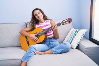 Evdeki kanepede oturmuş klasik gitar çalan genç ve güzel bir İspanyol kadın.