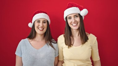 İzole edilmiş kırmızı arka planda kendine güvenen iki kadın Noel şapkası takıyor.