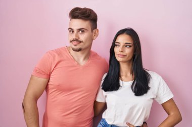 Young hispanic couple standing over pink background smiling looking to the side and staring away thinking. 