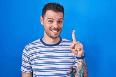 Young hispanic man standing over blue background showing and pointing up with finger number one while smiling confident and happy. 