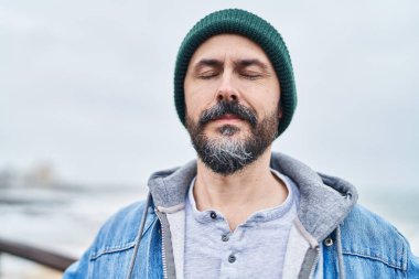 Genç kel adam deniz kenarında gözleri kapalı nefes alıyor.