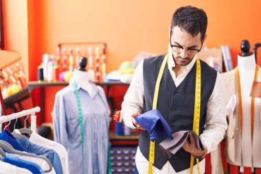 Young hispanic man tailor holding cloths at atelier