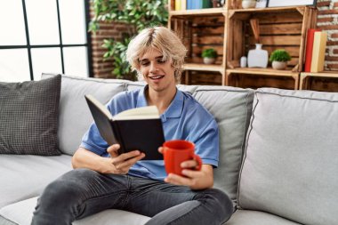 Genç sarışın adam kitap okuyor ve evde kahve içiyor.