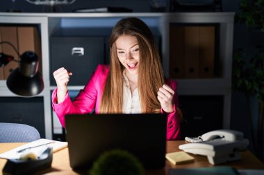 Geceleri ofiste çalışan beyaz bir kadın çok mutlu ve heyecanlı bir şekilde kolları havada, gülümseyen ve başarı için bağıran kazanan bir jest yapıyor. kutlama konsepti. 