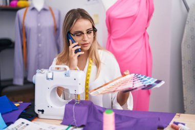 Young blonde woman tailor talking on smartphone choosing color at clothing shop