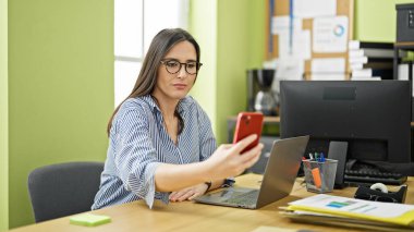 Genç, güzel İspanyol iş kadını ofisteki akıllı telefondan selfie çekiyor.