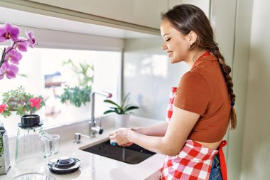 Young beautiful hispanic woman wearing apron washing glass at the kitchen