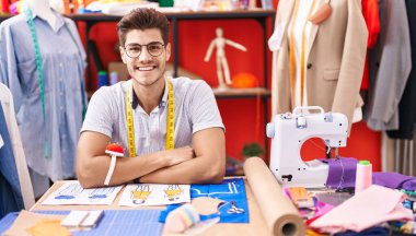 Young hispanic man tailor smiling confident sitting on table at atelier