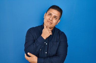 Mavi arka planda dikilen İspanyol genç adam elleri çenesinde soru sorarken, düşünceli bir ifade takınıyor. Düşünceli bir yüzle gülümsüyor. şüphe kavramı. 