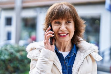 Orta yaşlı bir kadın gülümsüyor. Sokakta akıllı telefondan konuşuyor.
