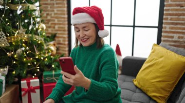 Beyaz saçlı, akıllı telefon kullanan olgun İspanyol kadın evdeki Noel ağacının yanında koltukta oturuyor.