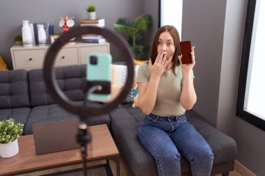 Güzel esmer kadın evde akıllı telefonuyla video kaydı yapıyor ağzını eliyle kapatıyor, şok geçiriyor ve hata yapmaktan korkuyor. şaşırmış ifade 