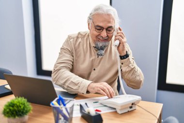 Middle age grey-haired man business worker talking on telephone using laptop at office